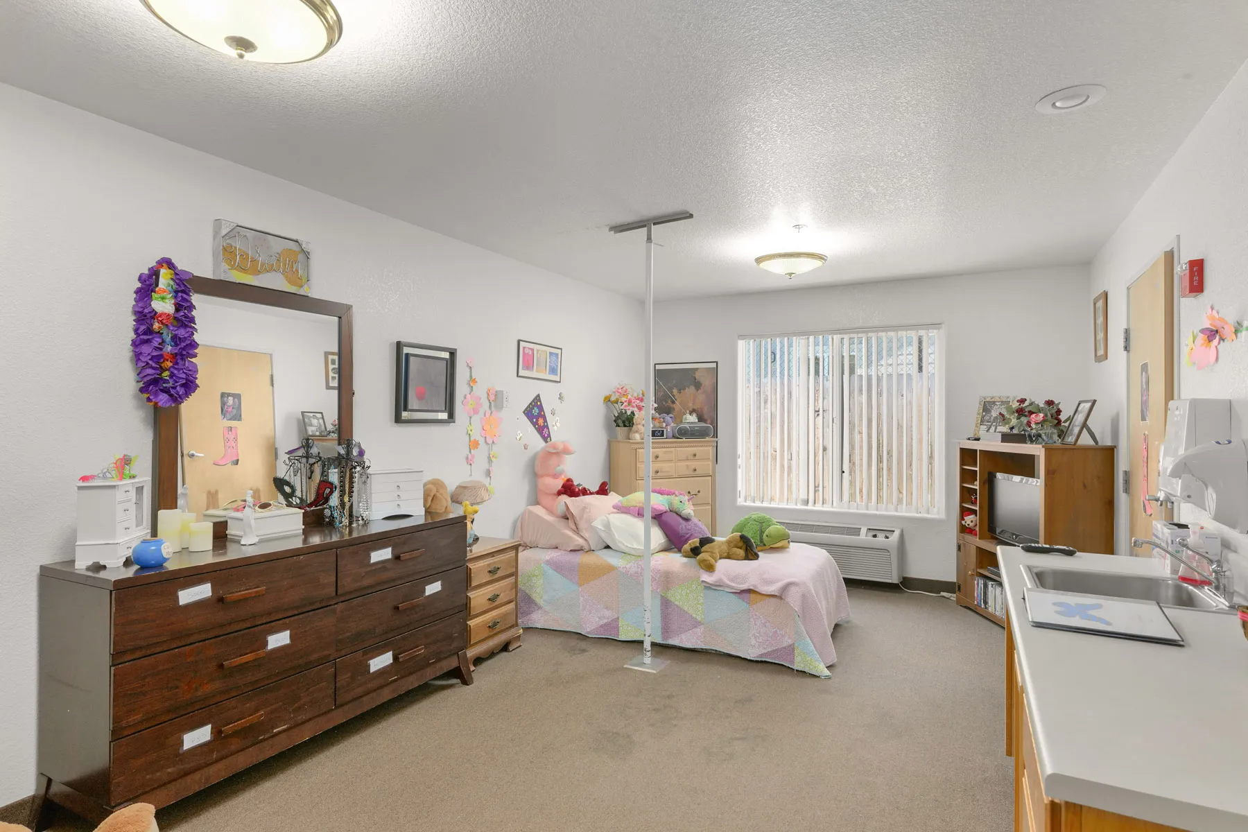 A cozy bedroom with a twin bed, colorful quilts, wooden furniture, and bright decor, featuring a window with blinds and a small kitchenette